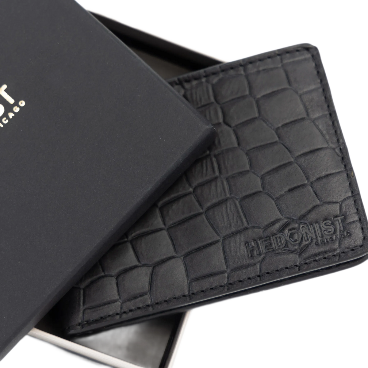 A black crocodile-embossed leather wallet shown partially open next to its black presentation box. The brand name 'HEDONIST Chicago' is debossed on the lower right corner of the wallet.