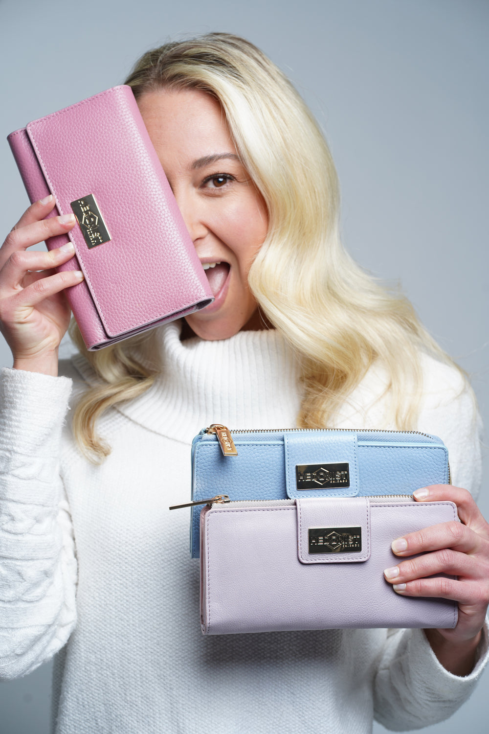 Woman holding three pink leather wallets with a visible brand logo against a gray background