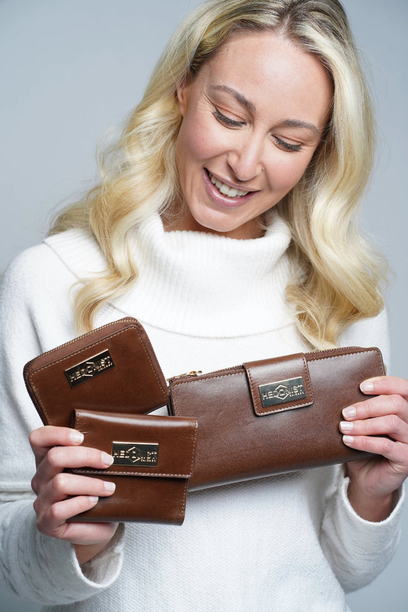 A smiling blonde woman in a white turtleneck sweater holds three brown leather wallets