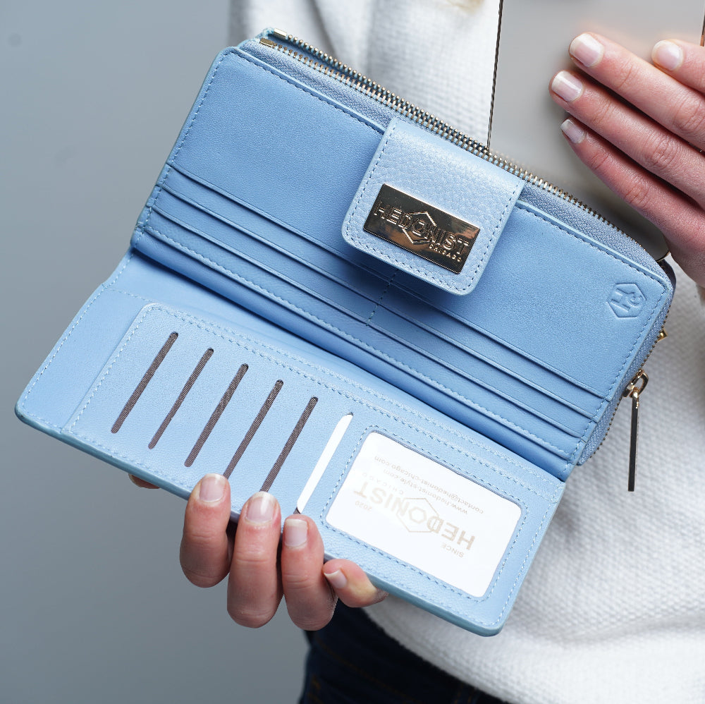 A light blue leather wallet shown open, held by a person. The wallet features multiple card slots, a zip-around closure, a gold-tone brand plaque on the front, and a white interior card labeled 'HEDONIST'.