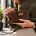 A woman in an olive green sweater holds a brown leather wallet with a gold-tone logo plate reading Hedonist Chicago.