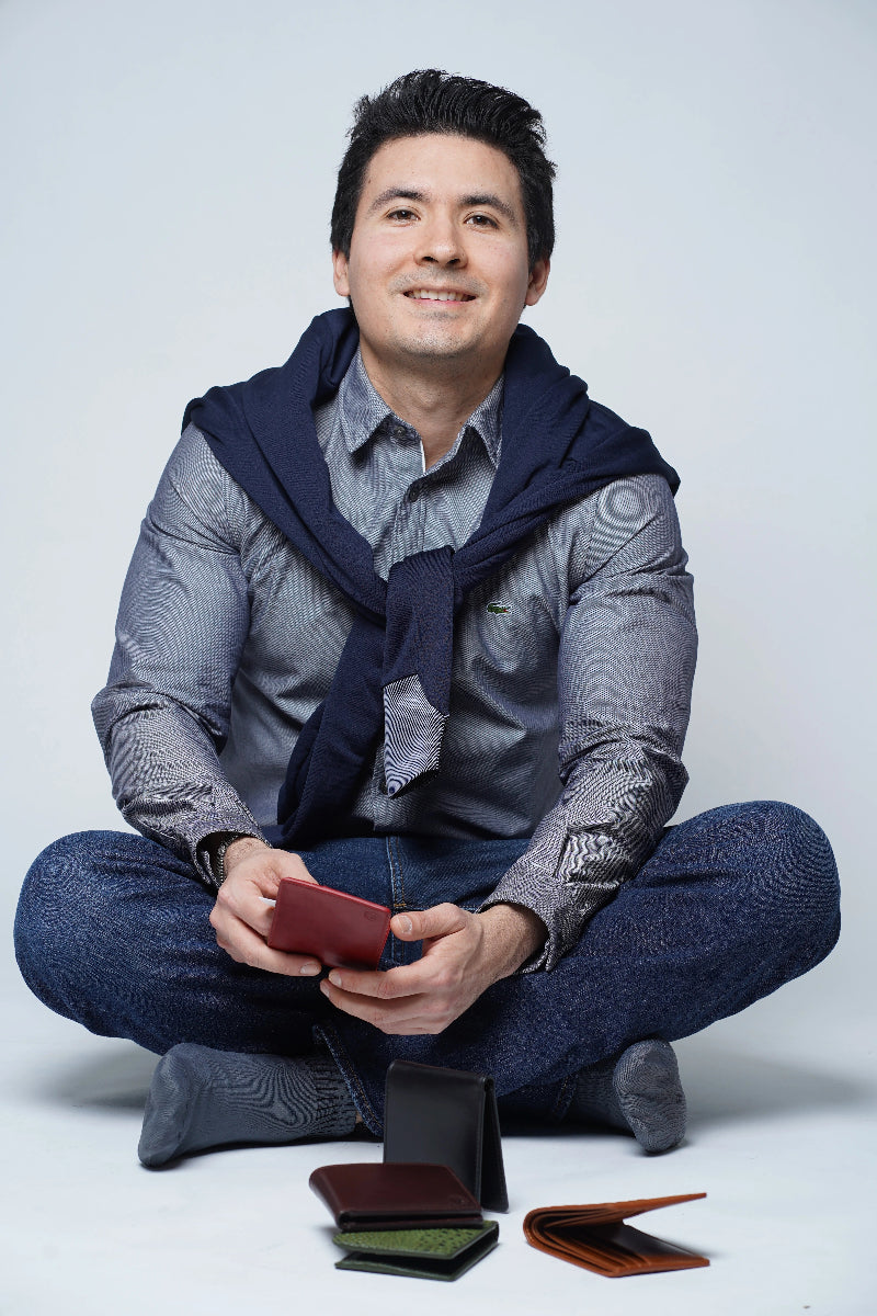 A man sitting cross-legged on a light gray studio floor. He is holding an open burgundy wallet, with several other wallets in different colors and materials arranged in front of him.