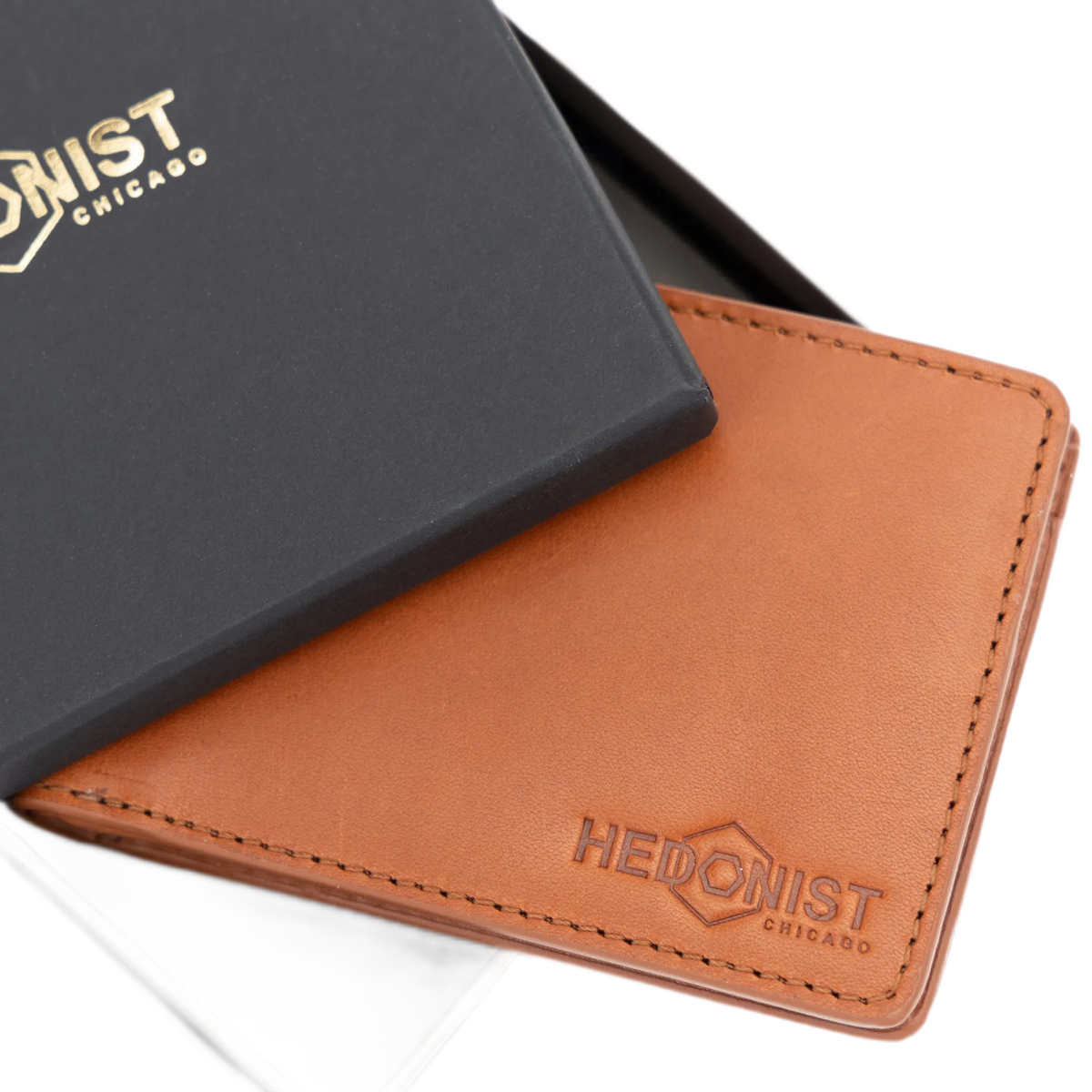 A tan leather bifold wallet with an embossed Hedonist Chicago logo, shown next to its black presentation box with gold lettering.