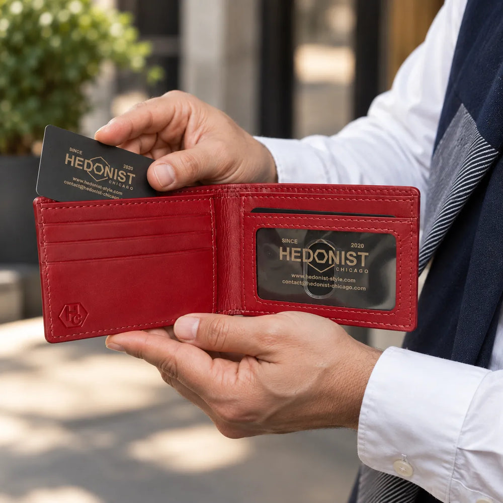 A red leather bifold wallet held open by a person, showing card slots, an ID window with a branded Hedonist card, and a small embossed logo on the lower left corner.