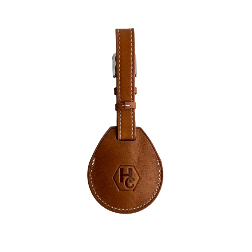 A brown leather AirTag Holder with a teardrop-shaped body, white contrast stitching, and a silver metal buckle, featuring an embossed 'HC' logo