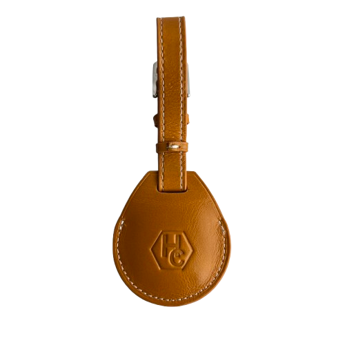 A tan leather AirTag Holder with a teardrop-shaped body, a matching leather strap, and a silver metal buckle, featuring an embossed 'HC' logo 