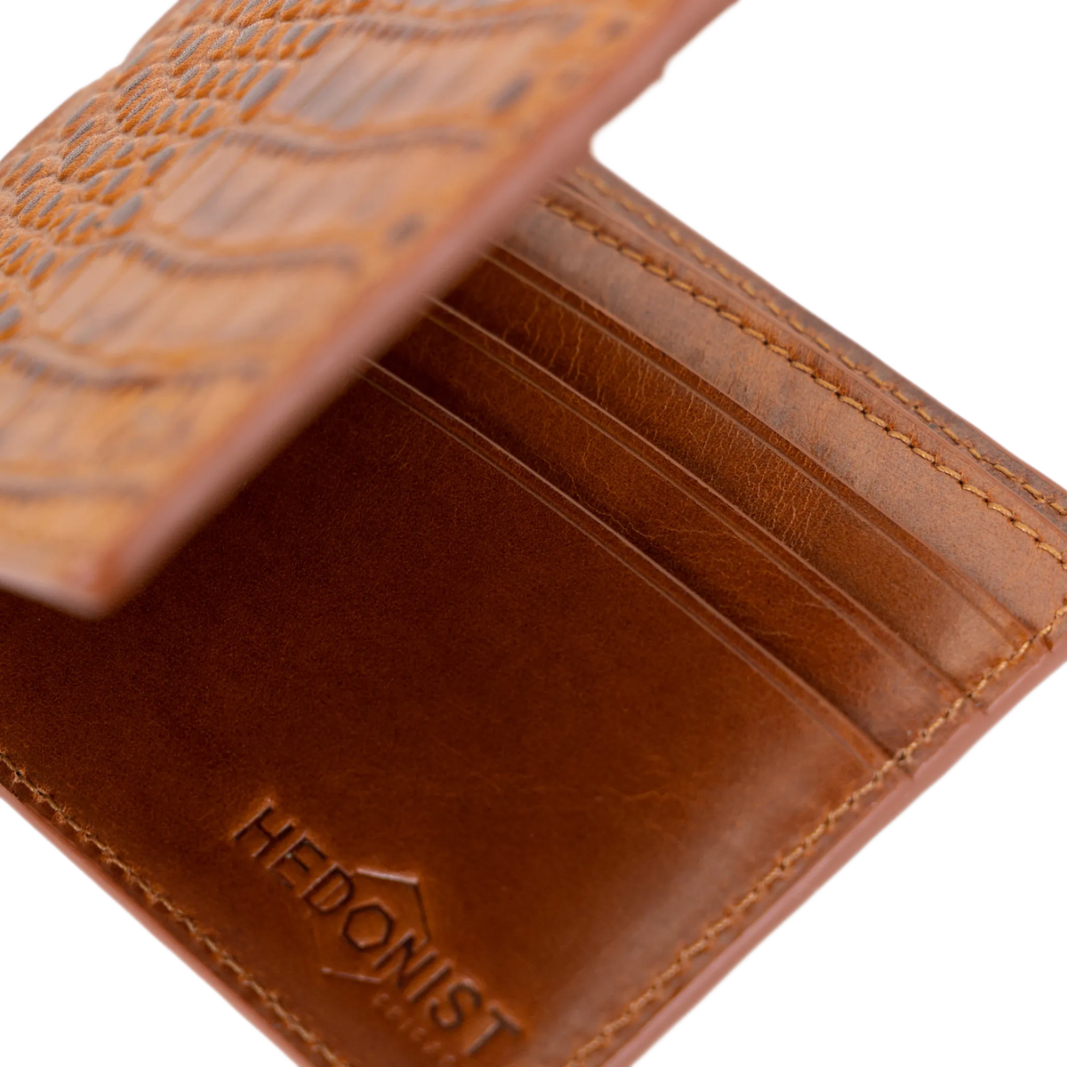 A close-up of a brown leather bifold wallet with an embossed crocodile pattern on the exterior and the brand name 'HEDONIST' debossed on the interior.