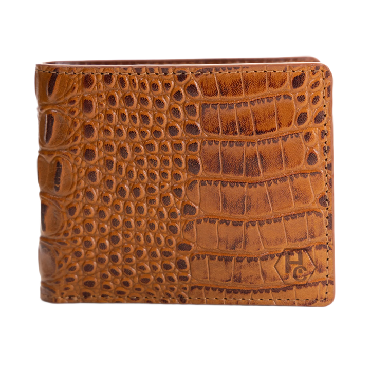 A brown crocodile-embossed leather bifold wallet with a stitched edge and a debossed monogram logo in the bottom right corner, shown against a plain white background.