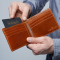 A person holds an open brown leather bifold wallet with multiple card slots and an embossed HEDONIST logo.