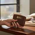 Hand holding a brown leather wallet with a visible brand logo on a wooden table.