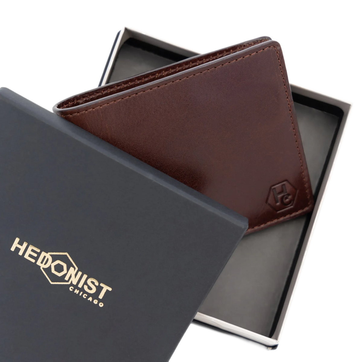 A dark brown leather bi-fold wallet with an embossed 'HC' logo, presented in an open black gift box next to its black branded outer lid which features the 'HEDONIST CHICAGO' logo in gold.