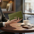 Woman holding the green croco-embossed leather trifold wallet with coffee on a table