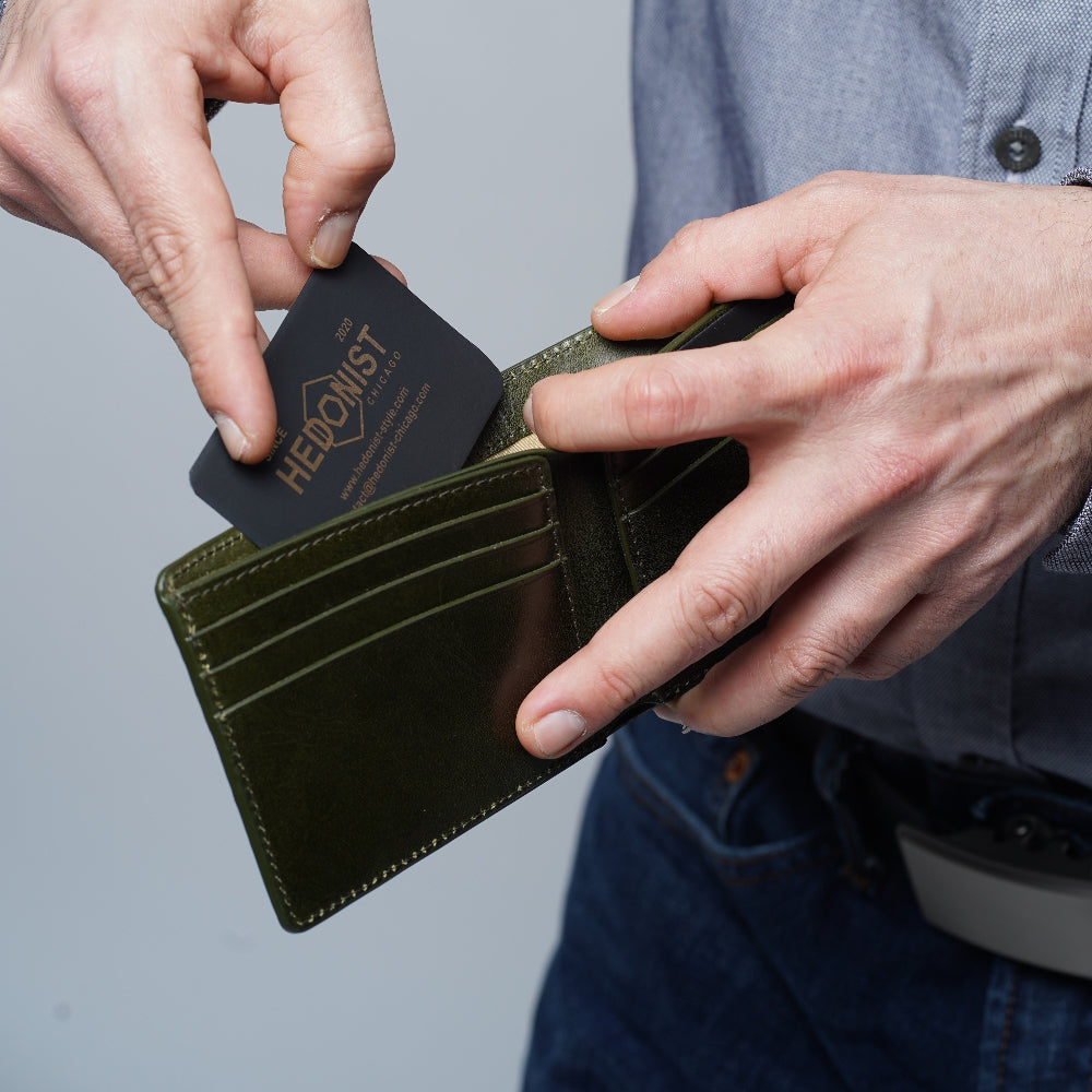 A close-up of a person's hands holding an olive green leather bifold wallet and inserting a black card. The person is wearing a blue button-down shirt and blue jeans against a neutral gray background.
