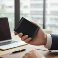 Hand holding a black minimalist men's wallet with a blurred office background