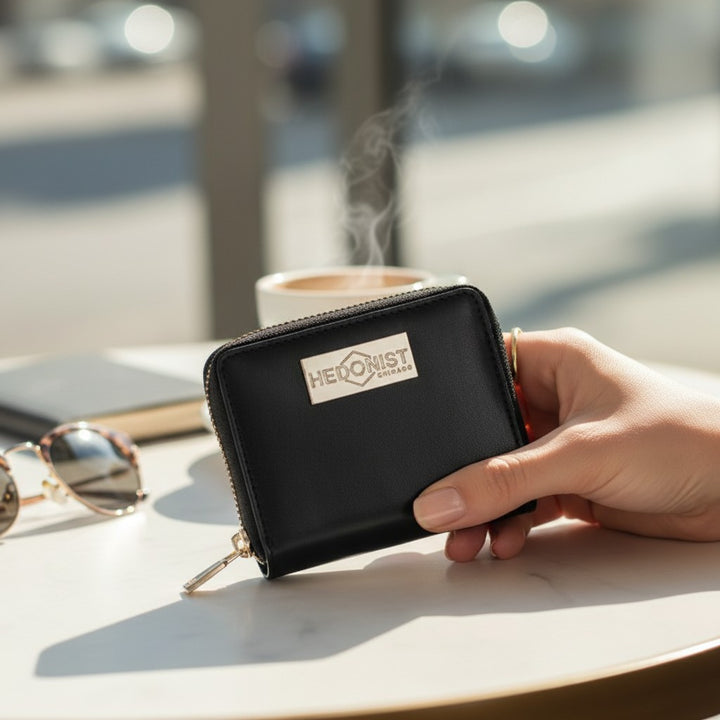 Hand holding a black wallet with 'Hedonist' branding on a table with sunglasses and a cup in the background.