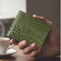 Hand holding a green textured wallet with a blurred background of a cup and saucer.