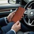 A man in a navy suit holding a brown handmade leather card holder wallet inside a car.