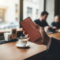 A hand holding a brown leather card holder wallet in a cafe setting.