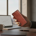 A hand holding a reddish-brown leather wallet or card holder in front of a blurred office 