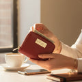 Women holding a red wallet with a Hedonist Chicago logo, sitting at a table with a cup of coffee and phone.