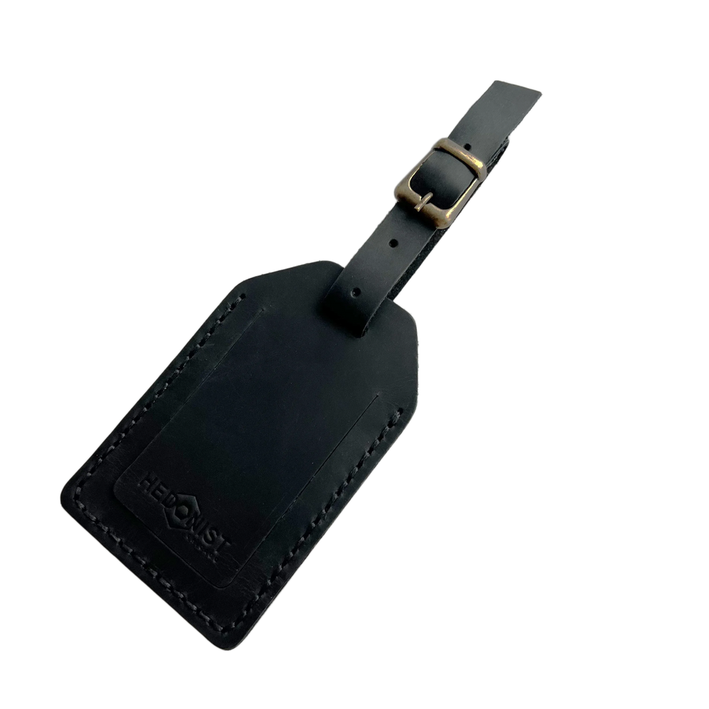 Black full-grain leather luggage tag with brass buckle and strap, embossed Hedonist Chicago logo on the front, shown on a light gray background.