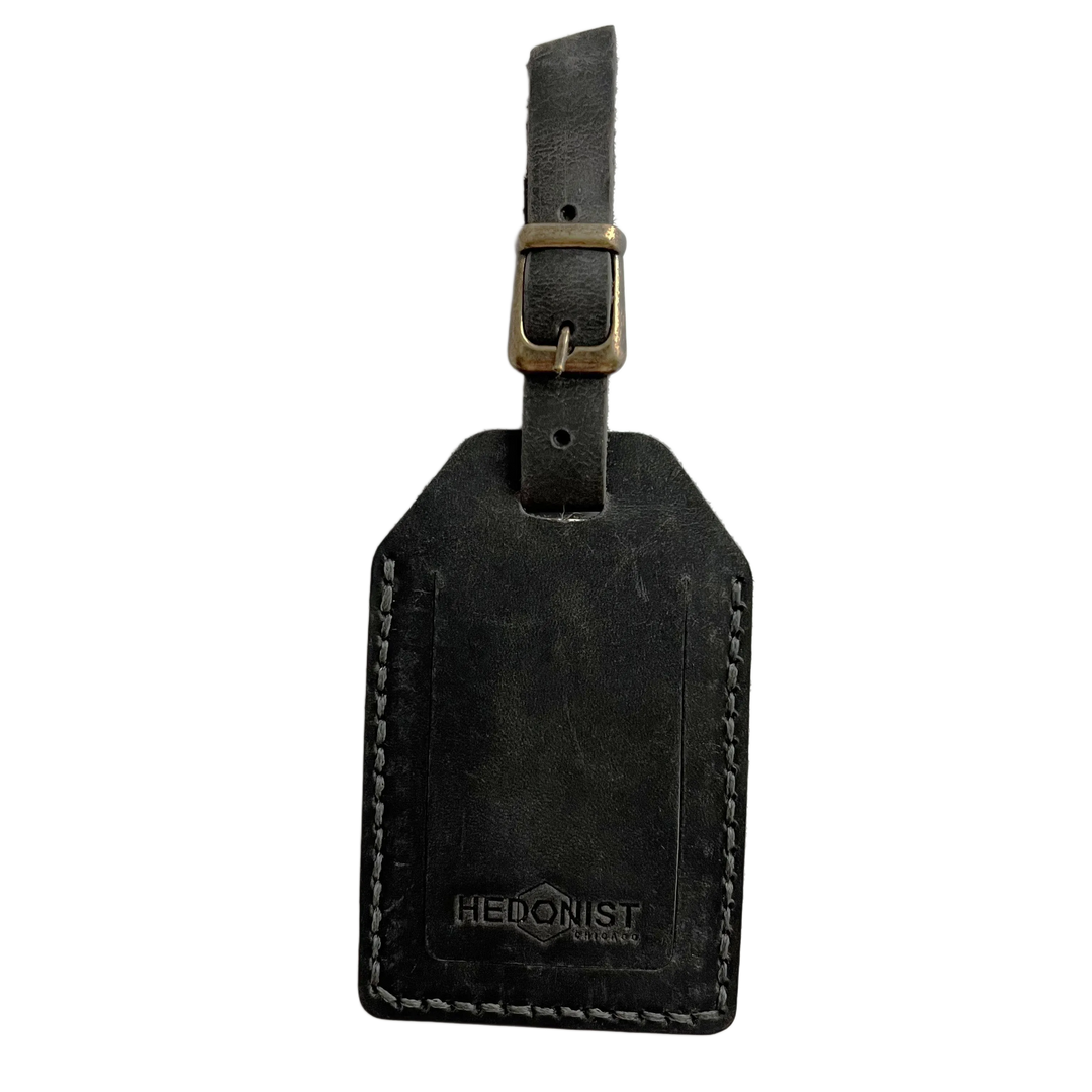 Black washed full-grain leather luggage tag with brass buckle strap, embossed Hedonist Chicago logo near the bottom, shown on a light background.