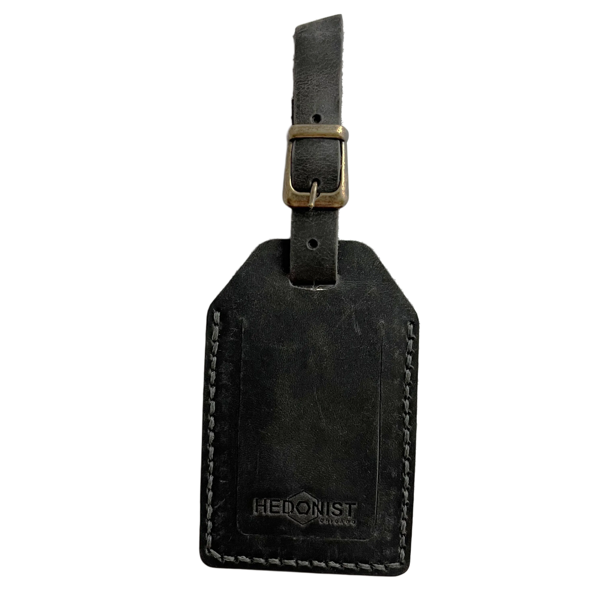 Black washed full-grain leather luggage tag with brass buckle strap, embossed Hedonist Chicago logo near the bottom, shown on a light background.