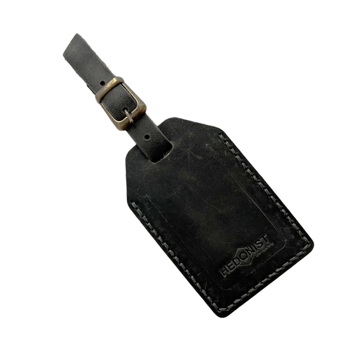 Black washed full-grain leather luggage tag with brass buckle, embossed Hedonist Chicago logo in the lower right corner, shown on a light gray background.