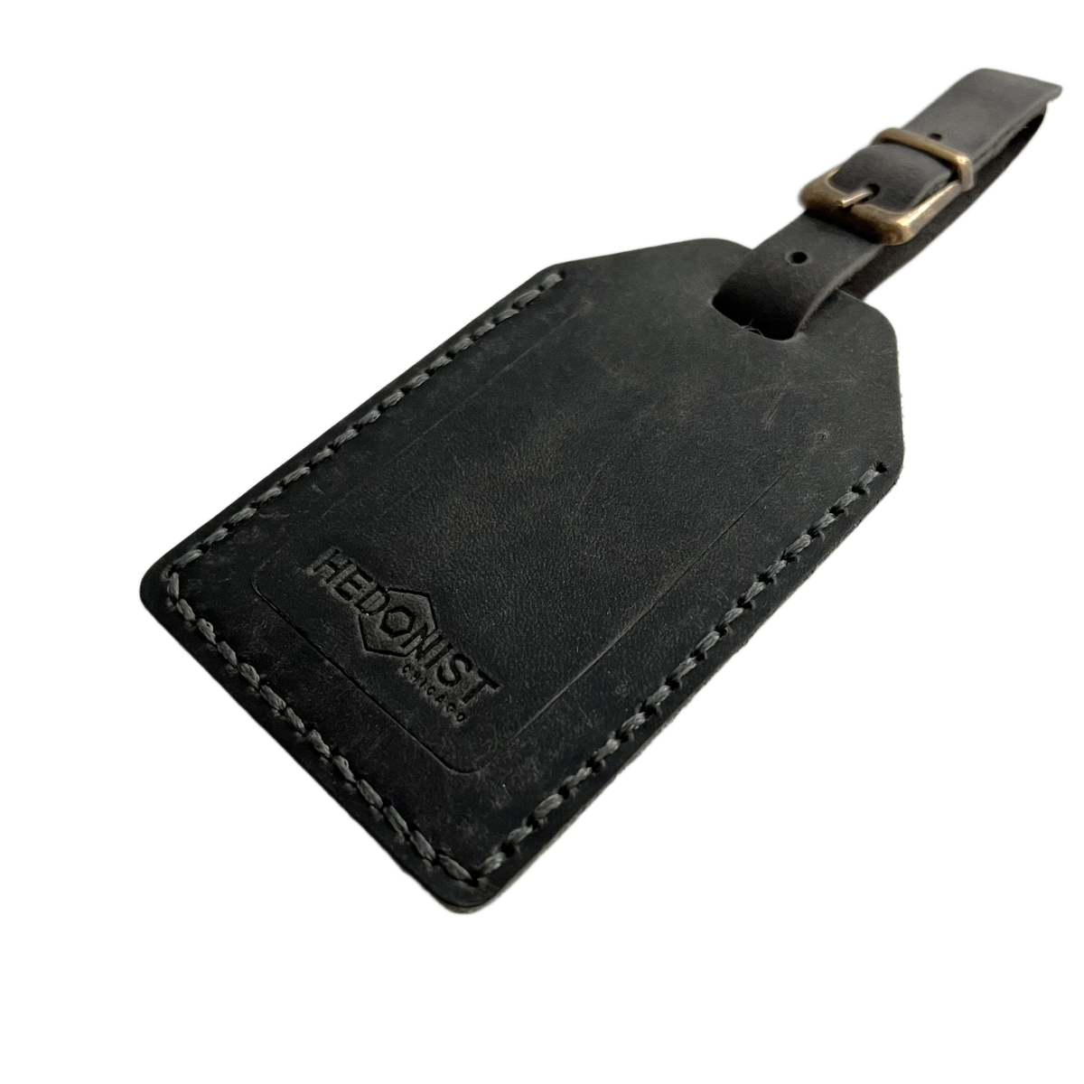 Black washed full-grain leather luggage tag with white contrast stitching and brass buckle, embossed Hedonist Chicago logo on the front, shown on a white background.