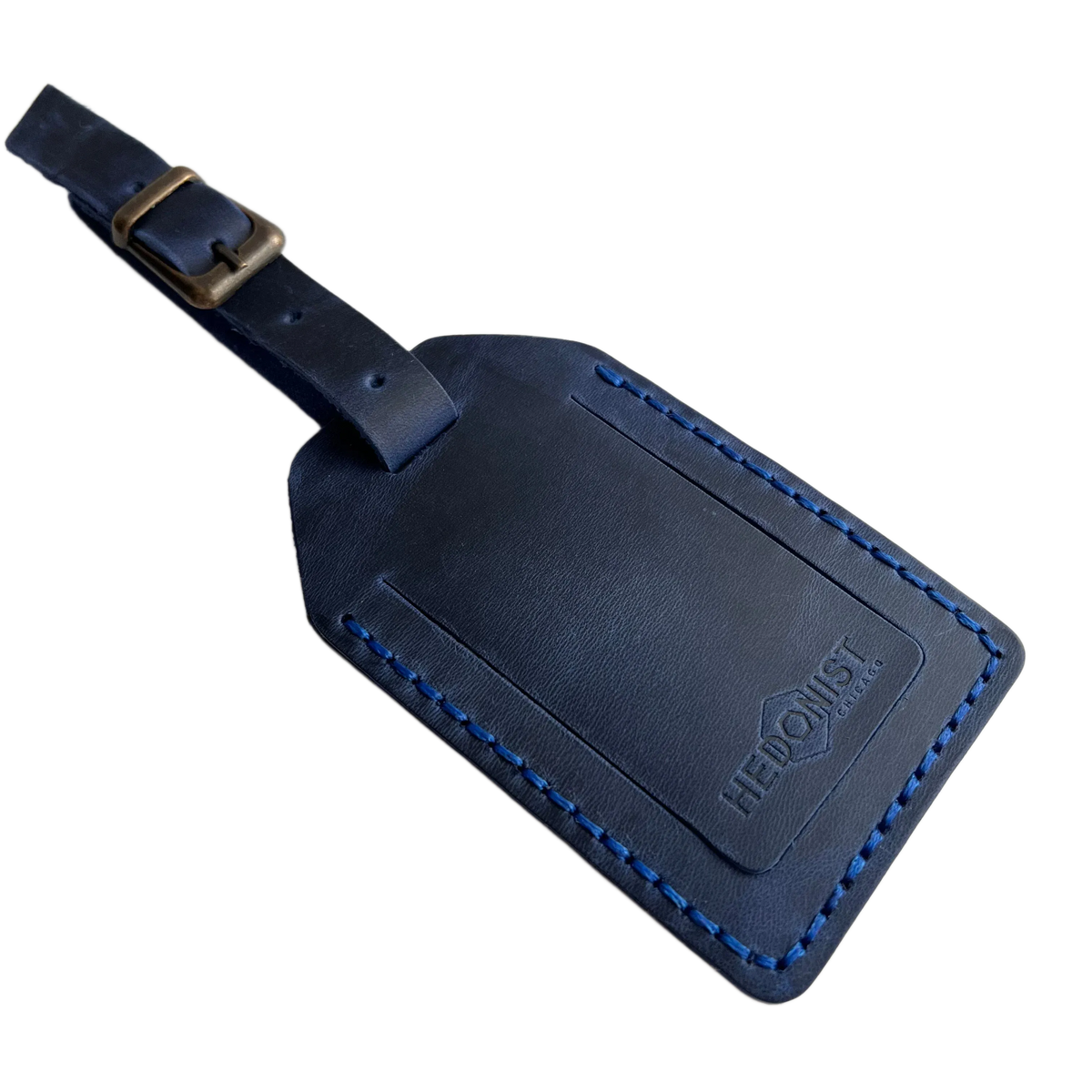 Navy blue full-grain leather luggage tag with blue contrast stitching and brass buckle strap, embossed Hedonist Chicago logo on the front, shown on an off-white background.