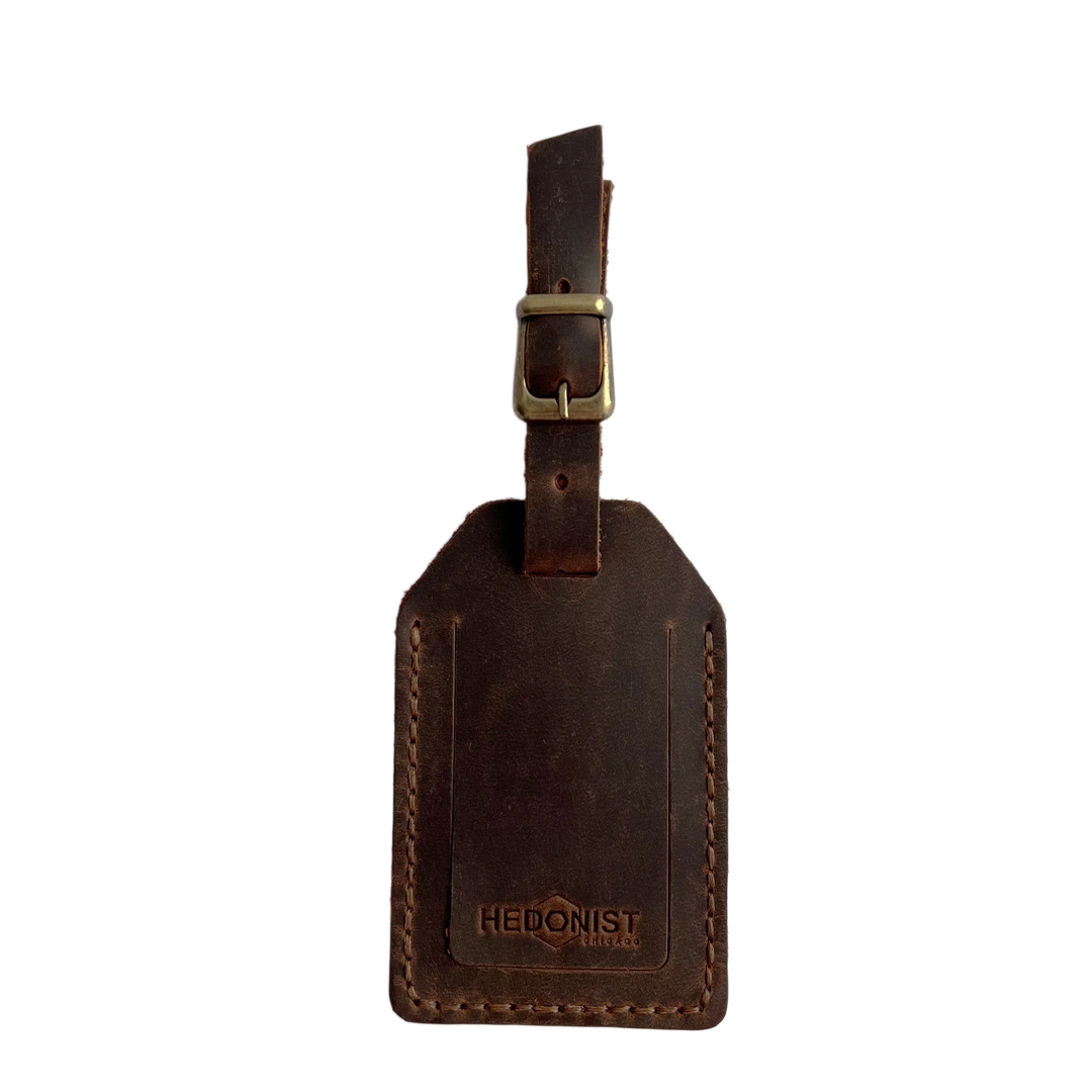 Brown full-grain leather luggage tag with brass buckle and strap, embossed Hedonist Chicago logo near the bottom, shown on a light gray background.