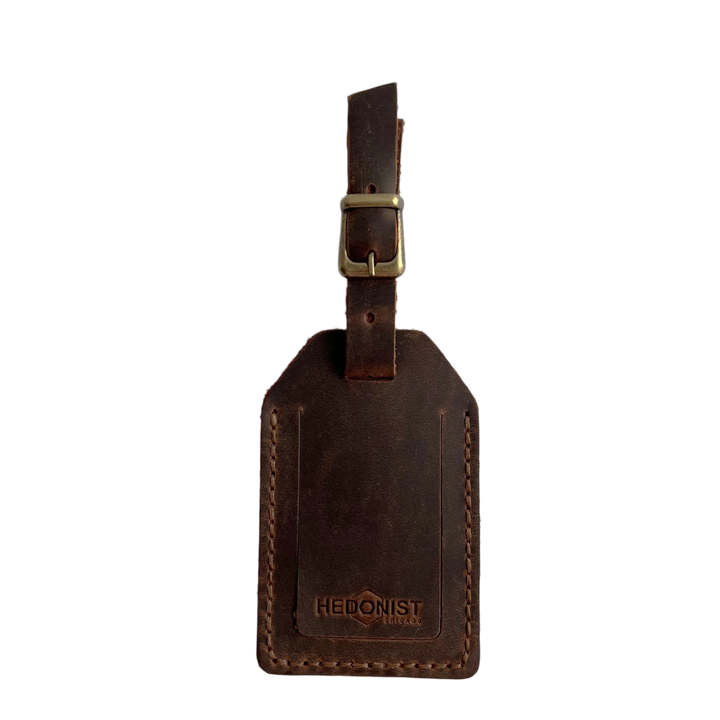 Brown full-grain leather luggage tag with brass buckle and strap, embossed Hedonist Chicago logo near the bottom, shown on a light gray background.