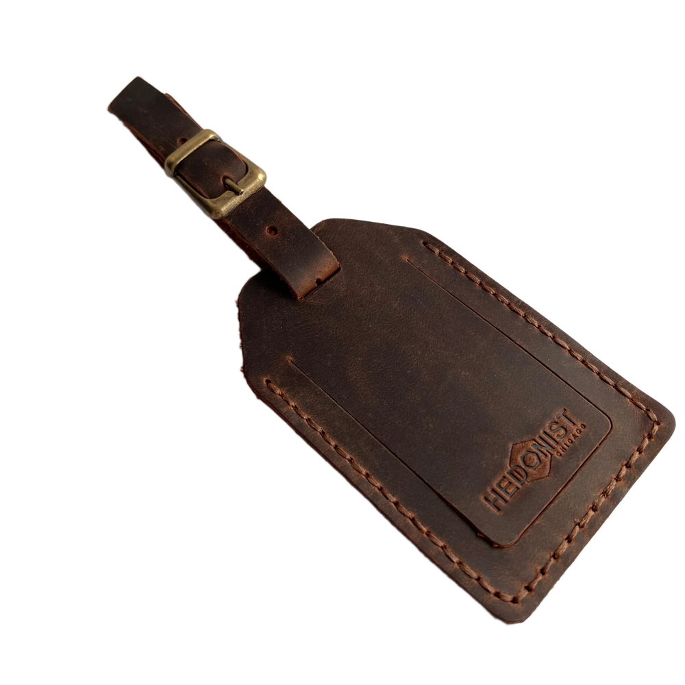 Brown full-grain leather luggage tag with brass buckle strap, embossed Hedonist Chicago logo in the lower right corner, shown on a light gray background.