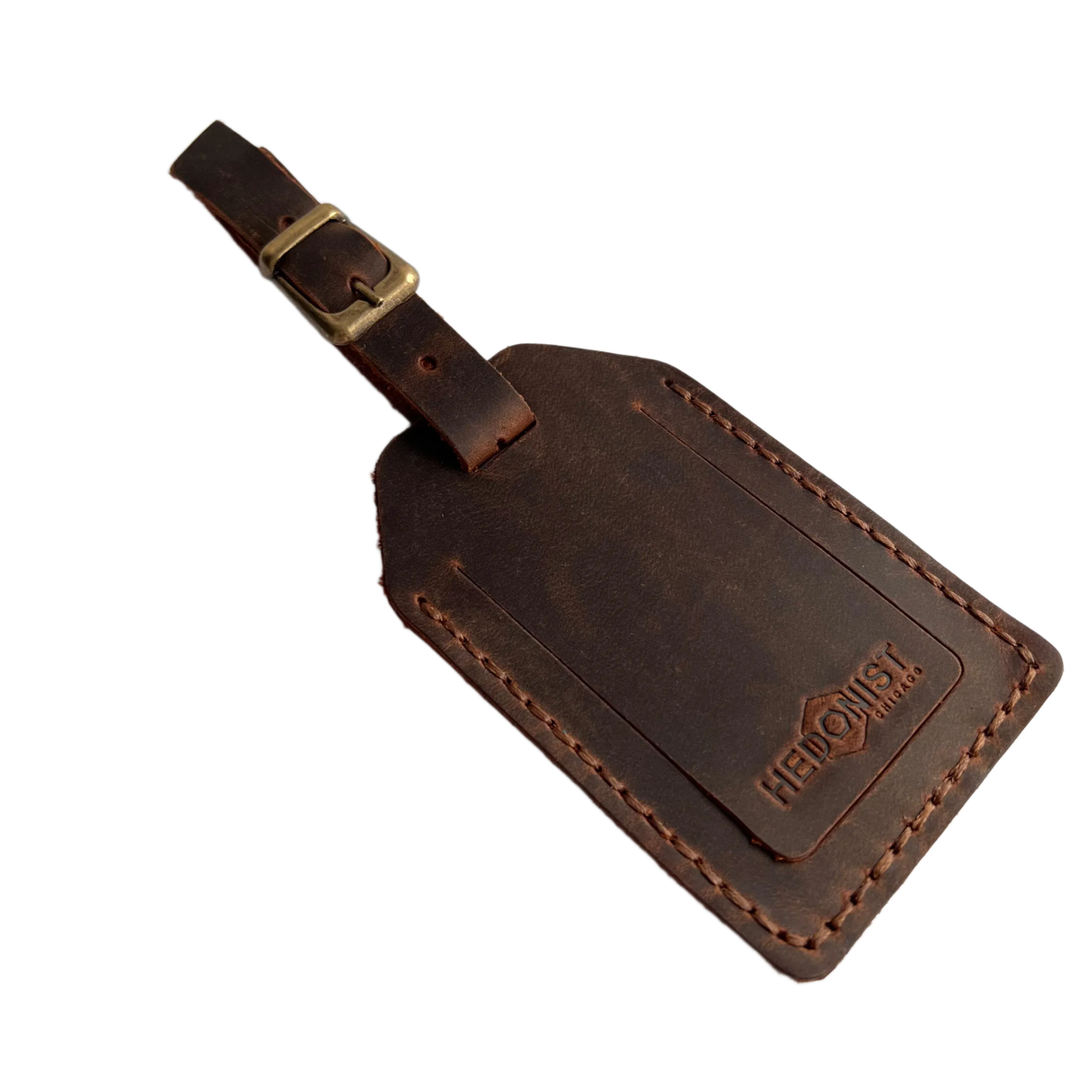 Brown full-grain leather luggage tag with brass buckle strap, embossed Hedonist Chicago logo in the lower right corner, shown on a light gray background.