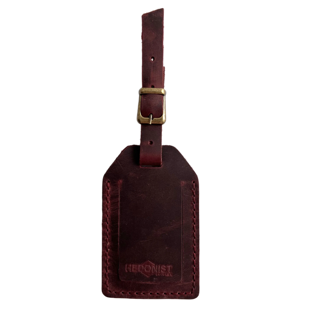 Burgundy full-grain leather luggage tag with brass buckle, embossed Hedonist Chicago logo near the bottom, shown on a white background.