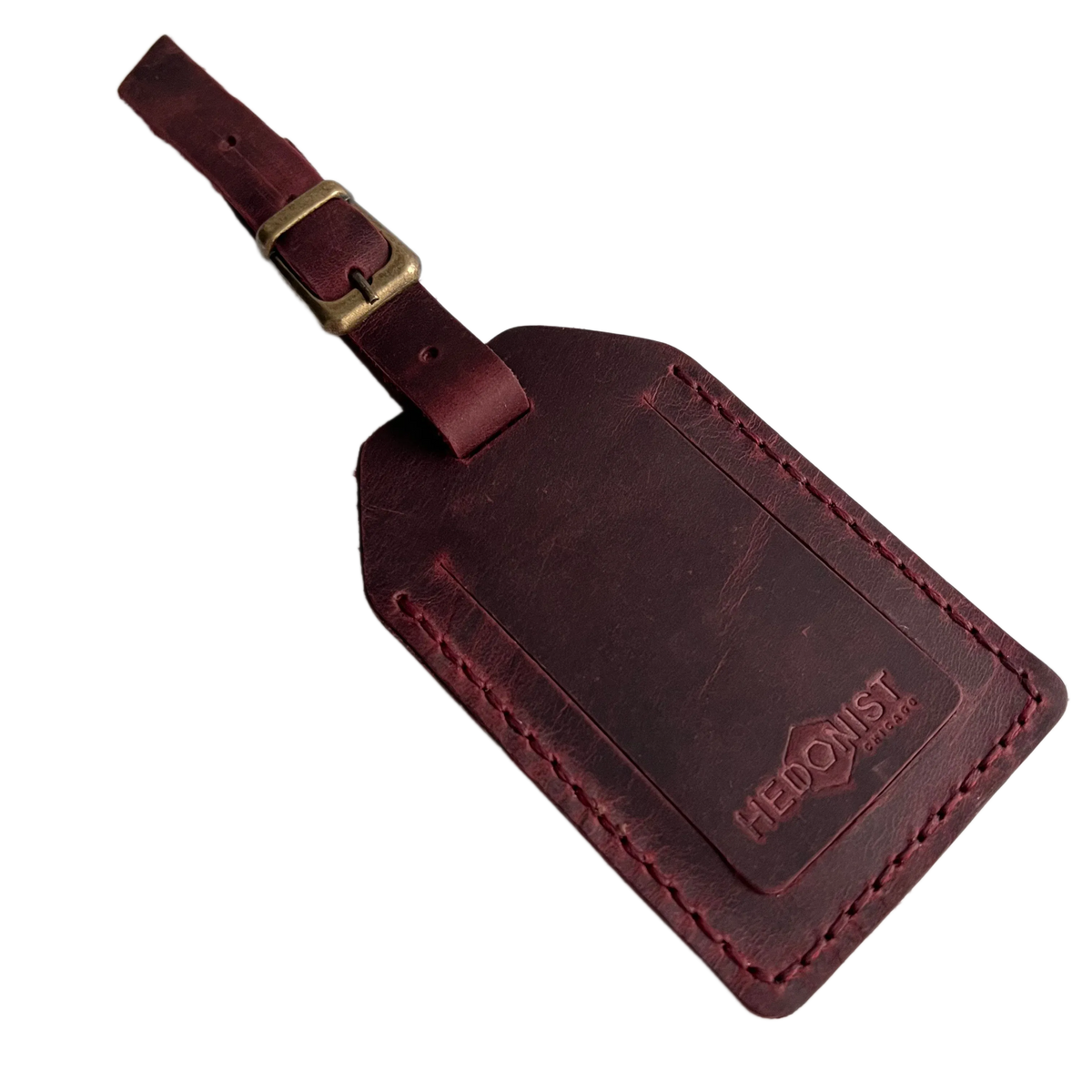 Burgundy full-grain leather luggage tag with matching leather strap and brass buckle, embossed Hedonist Chicago logo in the lower right corner, shown on a light gray background.