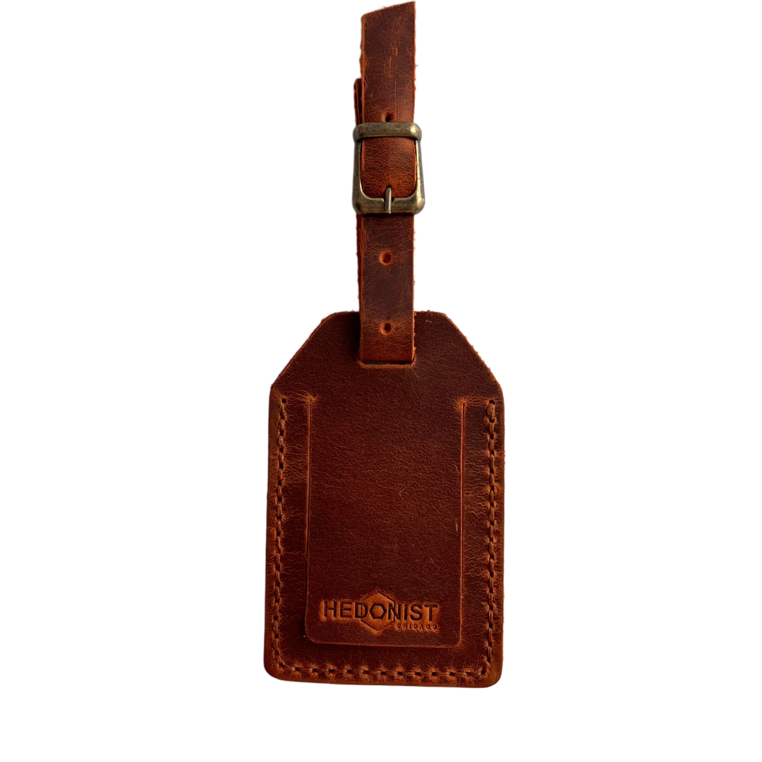 Cognac full-grain leather luggage tag with matching leather strap and brass buckle, embossed Hedonist Chicago logo on the front, shown on a light gray background.
