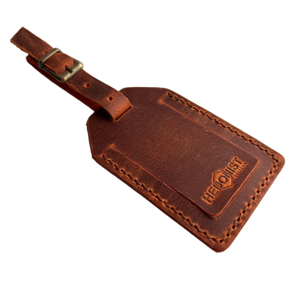 Cognac full-grain leather luggage tag with matching leather strap and brass buckle, embossed Hedonist Chicago logo on the front, shown on a light gray background.