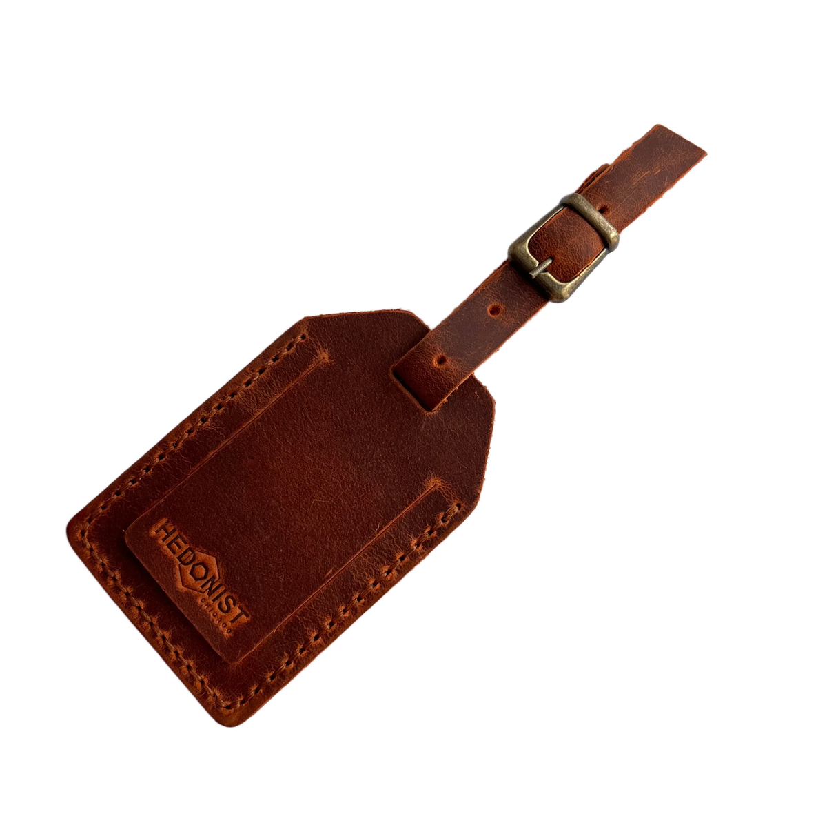 Cognac full-grain leather luggage tag with matching leather strap and brass buckle, embossed Hedonist Chicago logo on the front, shown on a light gray background.