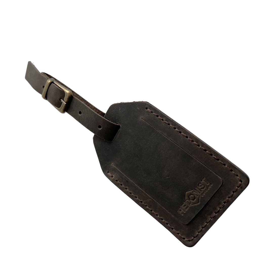 Dark brown full-grain leather luggage tag with matching leather strap and brass buckle, embossed Hedonist Chicago logo in the lower right corner, shown on a light gray background.