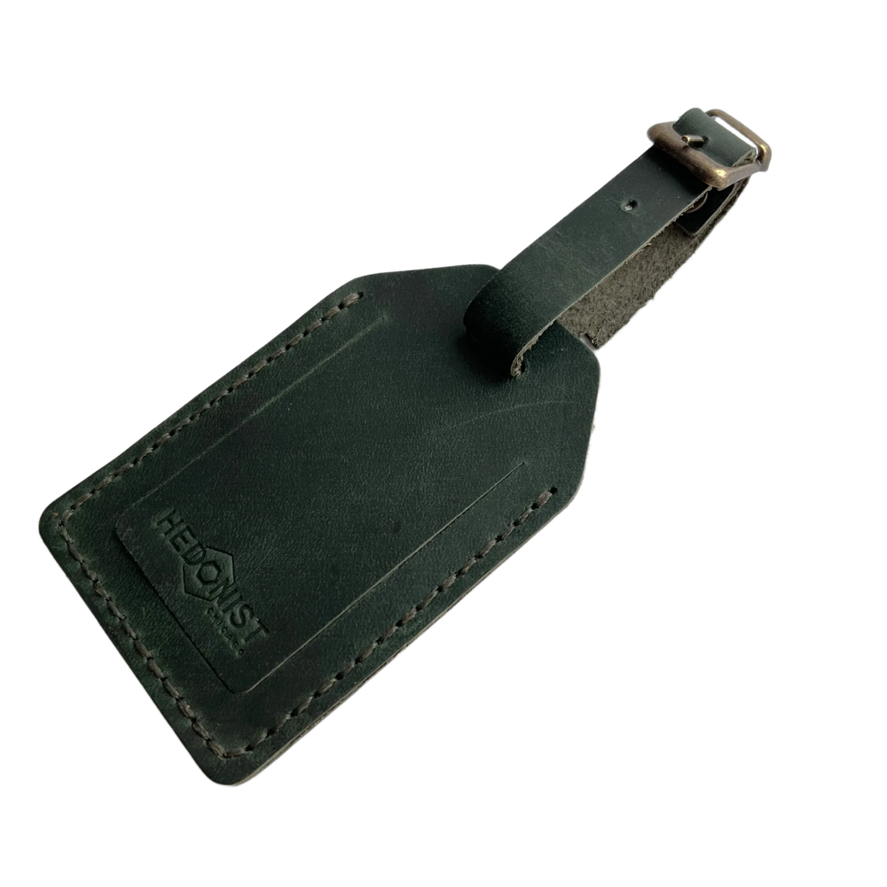 Green full-grain leather luggage tag with matching leather strap and brass buckle, embossed Hedonist Chicago logo on the front, shown on a light gray background.