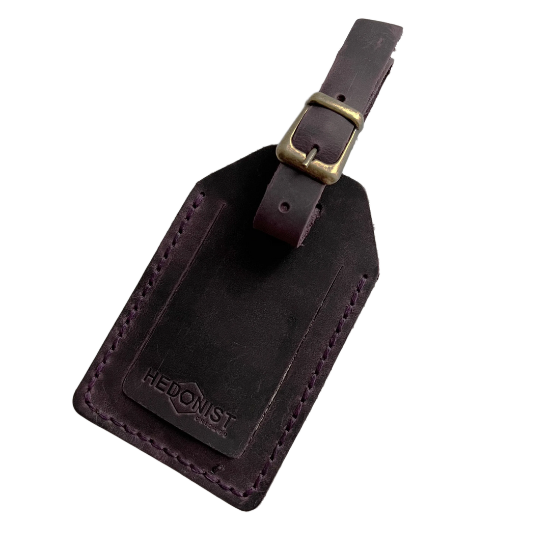 Plum full-grain leather luggage tag with matching leather strap and brass buckle, embossed Hedonist Chicago logo on a stitched leather patch.