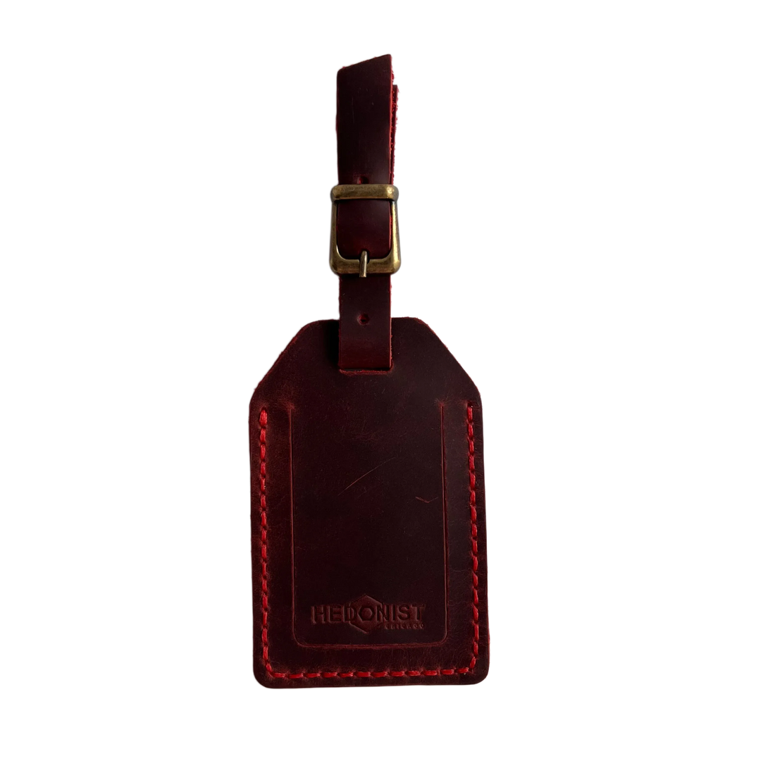 Red full-grain leather luggage tag with brass buckle and strap, embossed Hedonist Chicago logo on the front, shown on a white background.