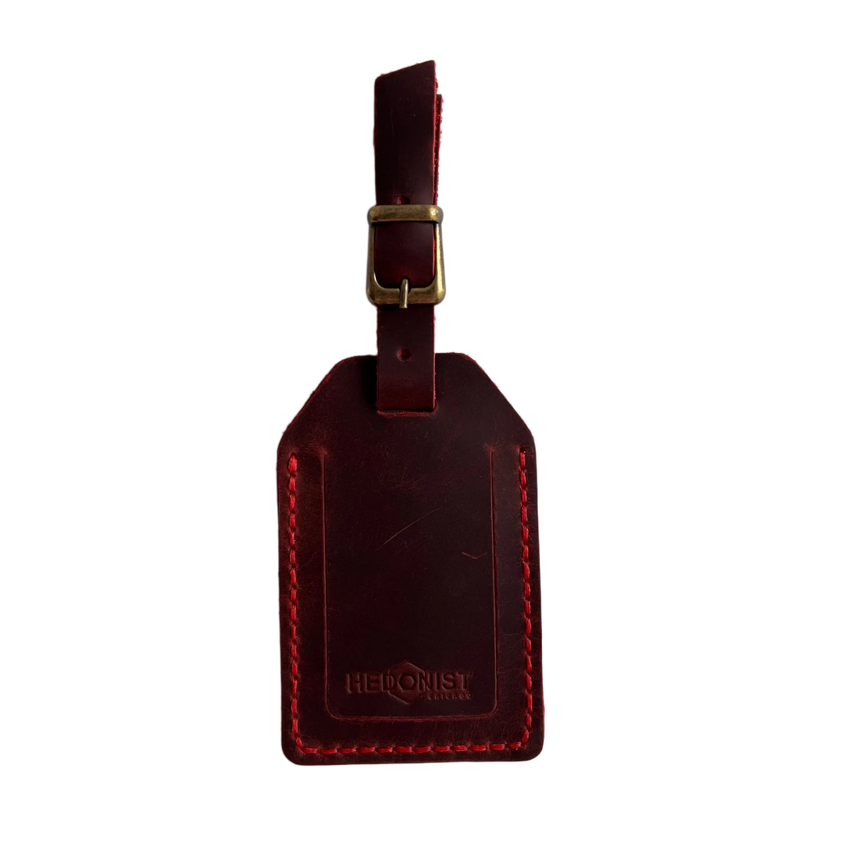 Red full-grain leather luggage tag with brass buckle and strap, embossed Hedonist Chicago logo on the front, shown on a white background.