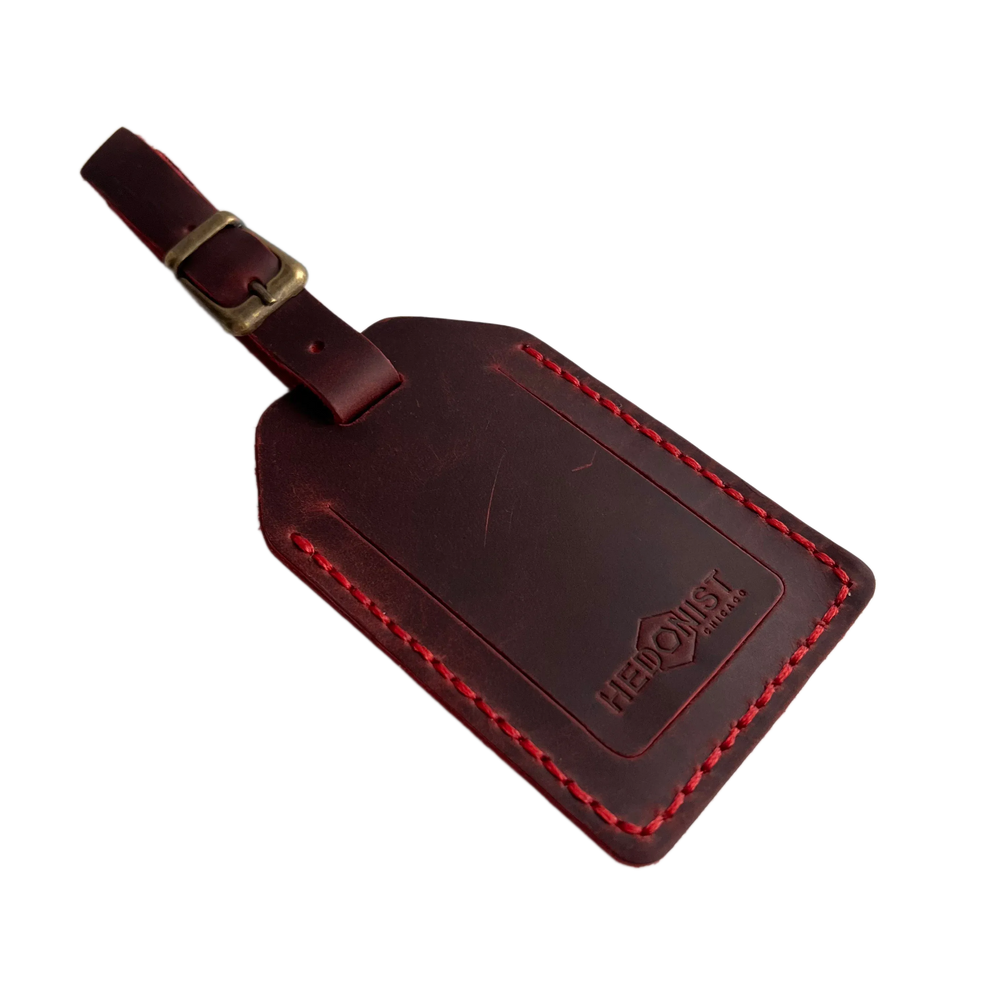 Red full-grain leather luggage tag with red stitching and brass buckle strap, embossed Hedonist Chicago logo on the front, shown on a white background.
