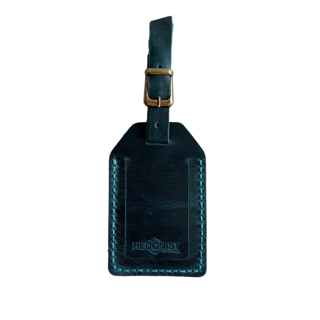 Teal full-grain leather luggage tag with matching leather strap and brass buckle, teal stitching, embossed Hedonist Chicago logo near the bottom, shown on a white background.
