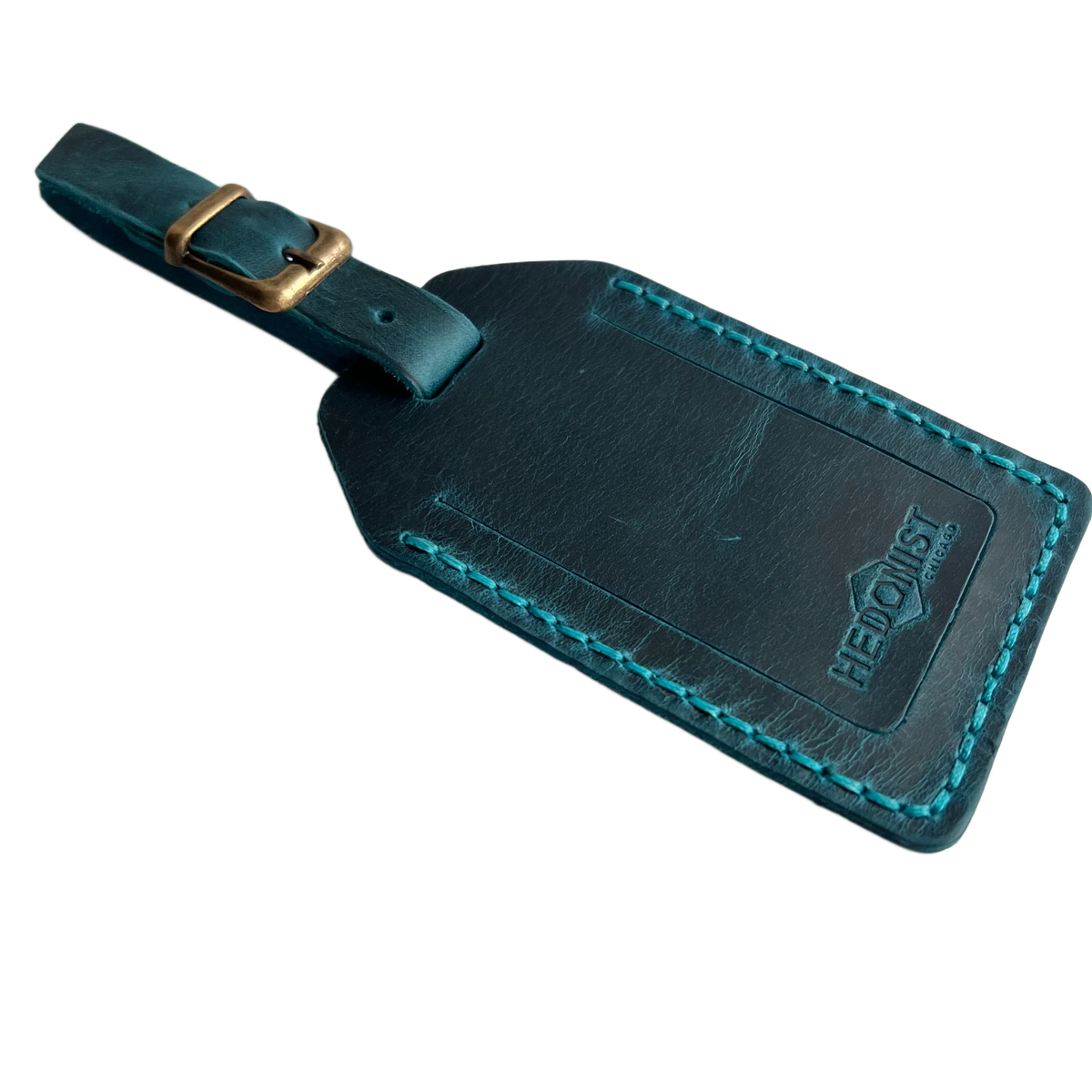 Teal full-grain leather luggage tag with brass buckle and matching teal stitching, embossed Hedonist Chicago logo on the front, shown on a light gray background.